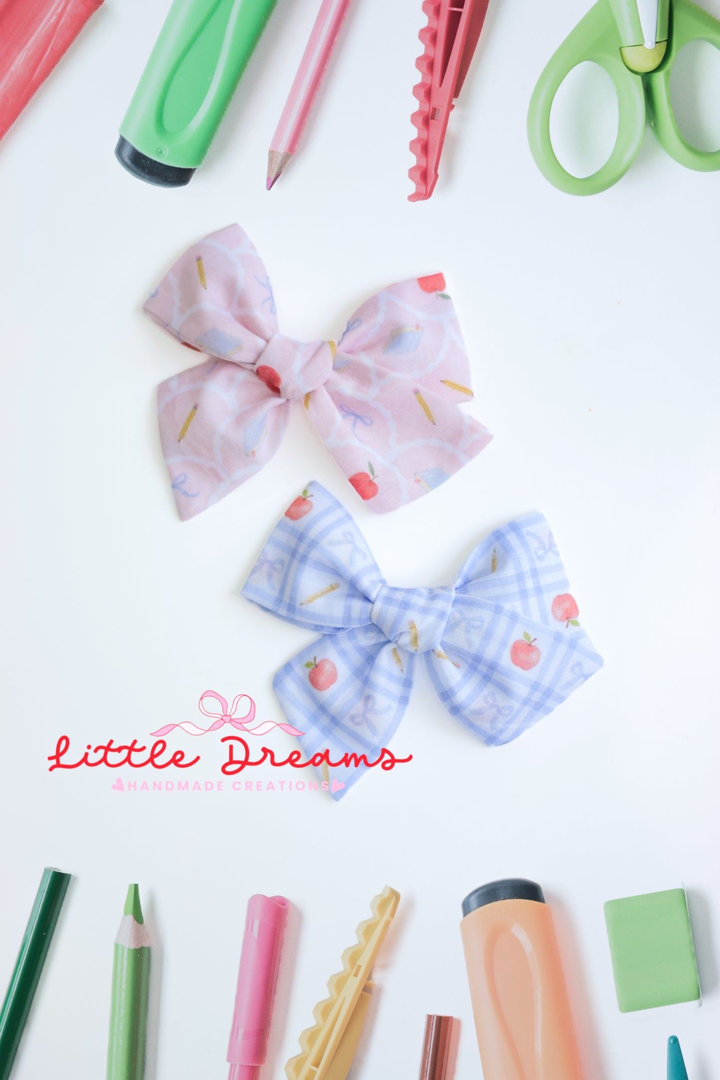 Two fabric hair bows displayed on a white background, surrounded by colorful stationery props. The top bow is light pink with a subtle pattern of notebooks, apples, pencils and bows, and the bottom bow is light blue with a gingham pattern and apples, pencils and bows.