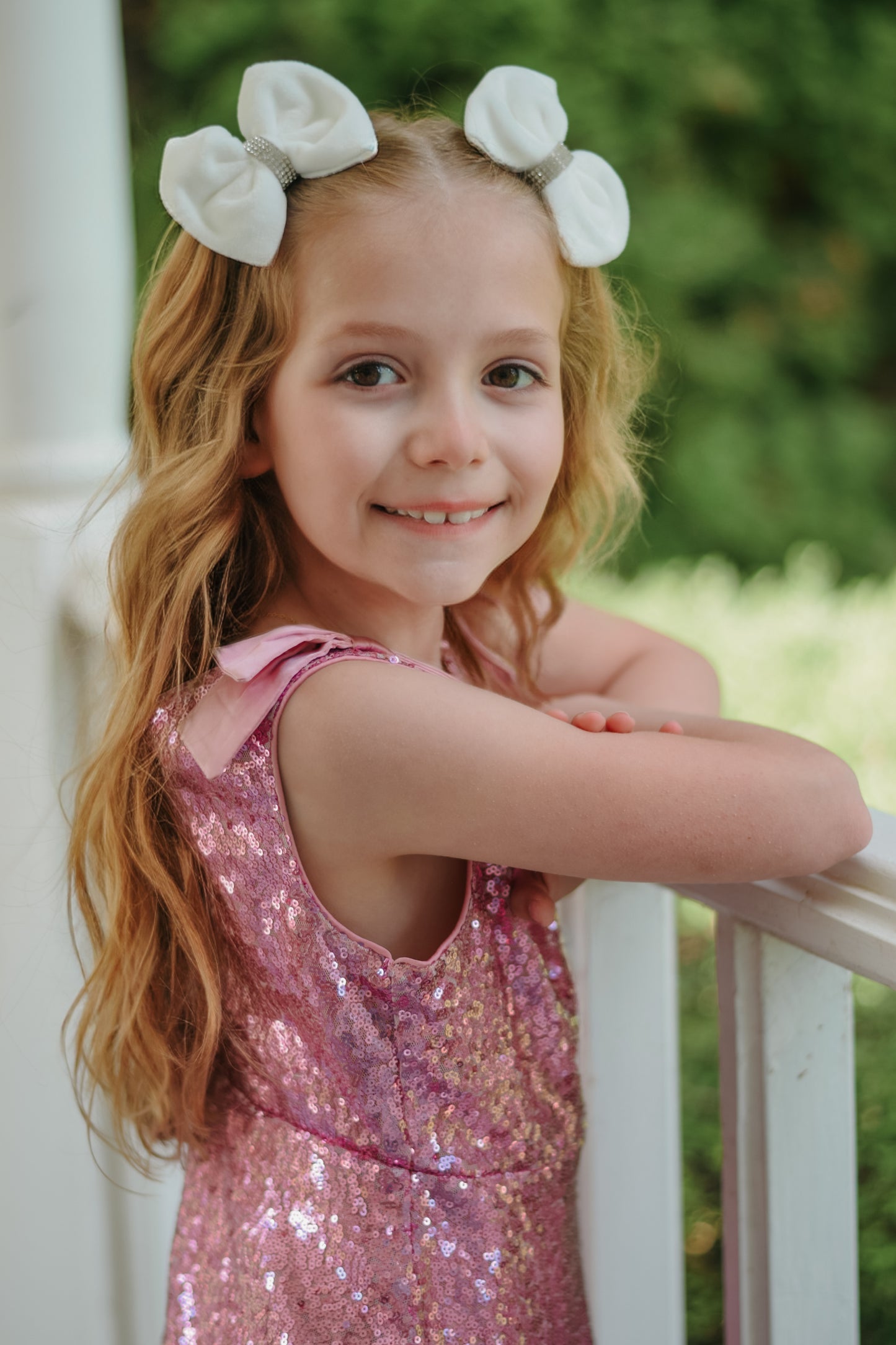 A smiling young girl with long, wavy blonde hair leans on a white porch railing. She is wearing a pink sequined dress with bow shoulder straps and two white velvet hair bows featuring rhinestone centers.
