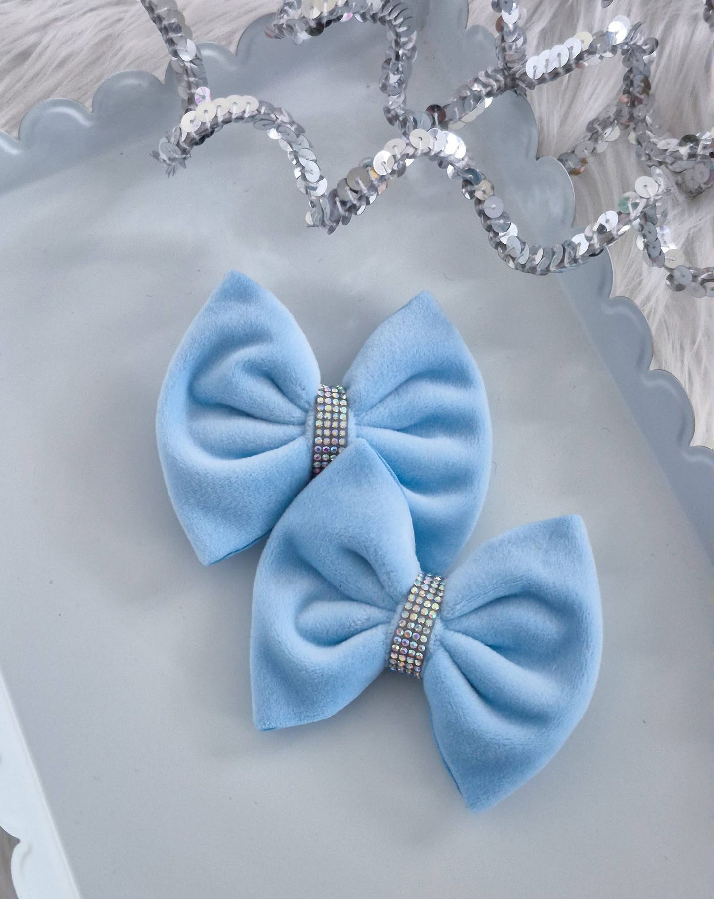 Two light blue velvet hair bows with rhinestone-encrusted center bands, displayed on a white scalloped tray with a silver sequin garland in the background.