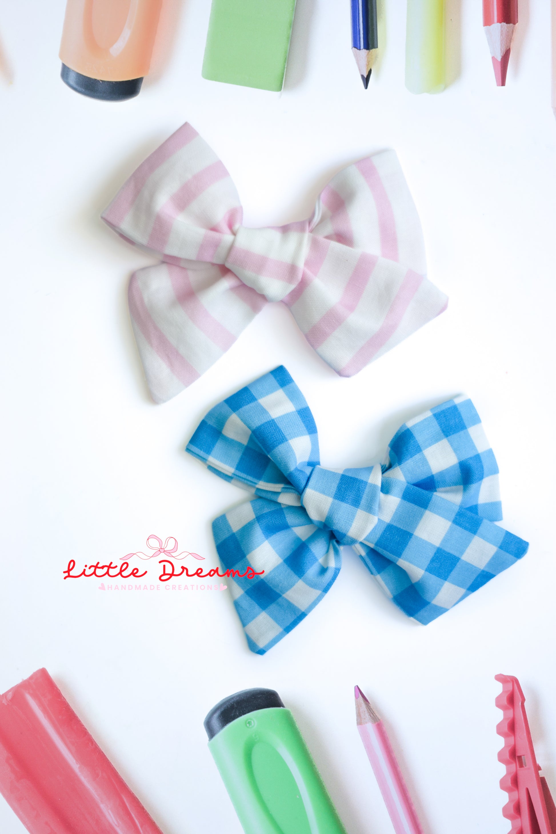Two fabric hair bows displayed on a white background with school supplies around them. The top bow has a pink and white striped pattern, and the bottom bow has a blue and white gingham pattern.
