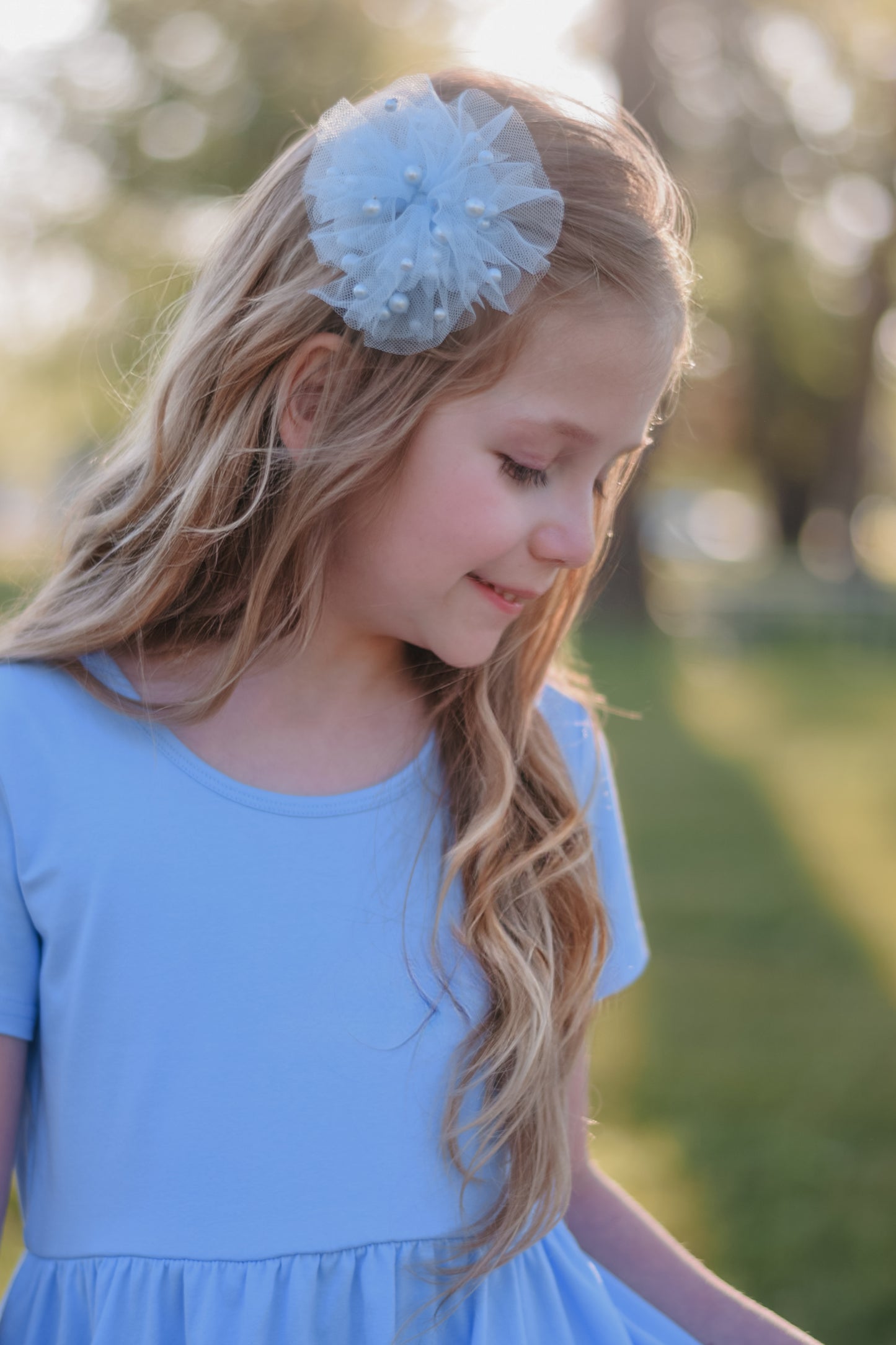 A young girl with long, wavy blonde hair looking down and smiling softly outdoors. She is wearing a light blue dress and a matching light blue tulle hair bow embellished with small pearls.