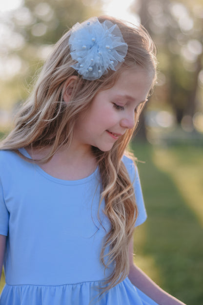 A young girl with long, wavy blonde hair looking down and smiling softly outdoors. She is wearing a light blue dress and a matching light blue tulle hair bow embellished with small pearls.