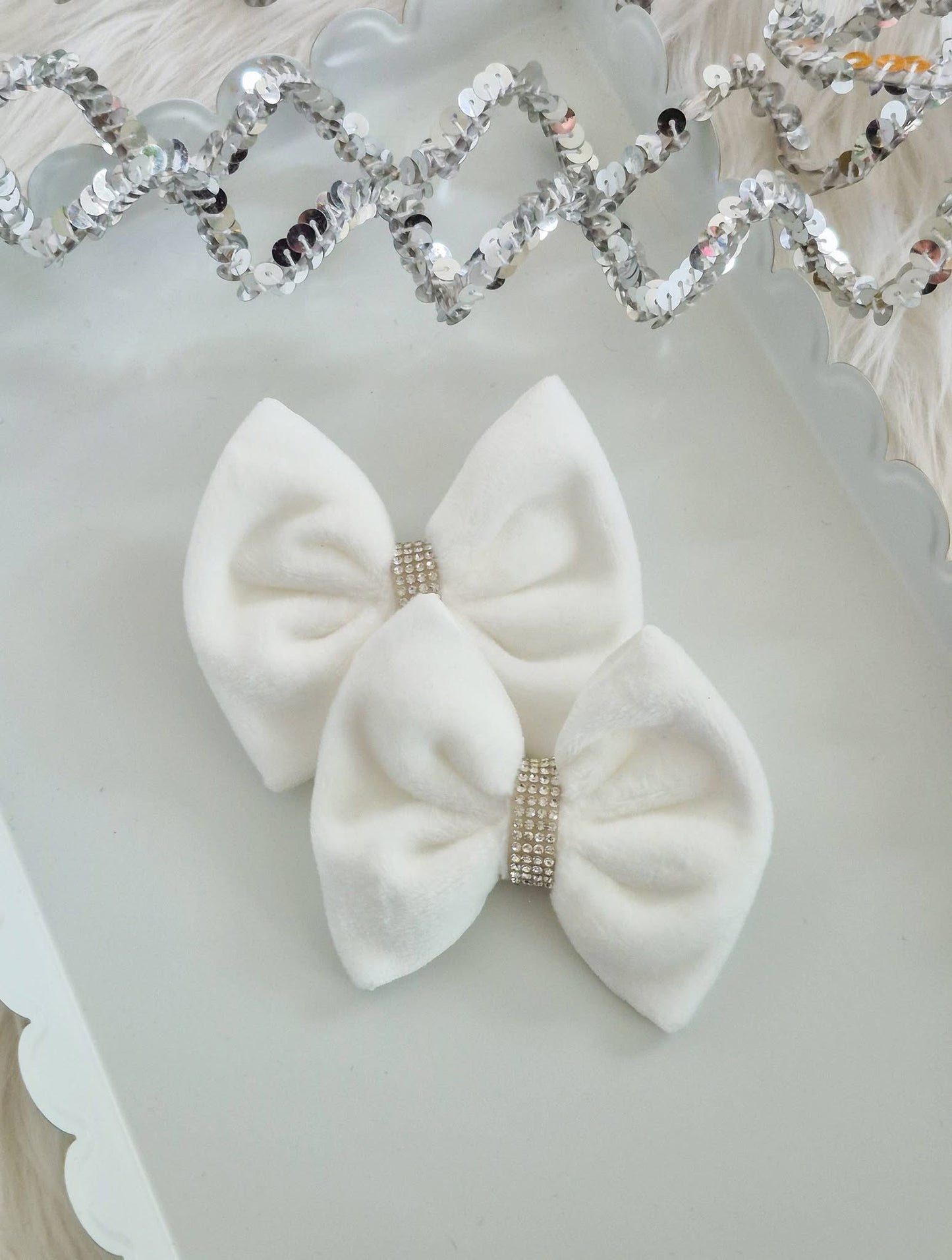 Two white velvet hair bows with rhinestone-encrusted center bands, displayed on a white scalloped tray with a silver sequin garland in the background.