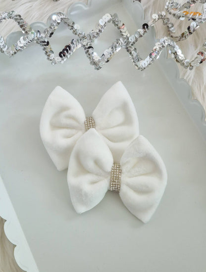 Two white velvet hair bows with rhinestone-encrusted center bands, displayed on a white scalloped tray with a silver sequin garland in the background.