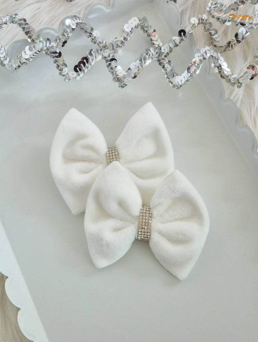 Two white velvet hair bows with rhinestone-encrusted center bands, displayed on a white scalloped tray with a silver sequin garland in the background.