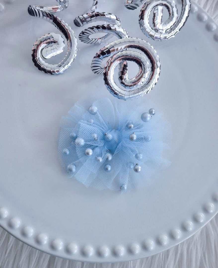 A light blue tulle hair bow embellished with scattered pearl beads, displayed on a white beaded-edge plate with metallic silver spiral decorations in the background.