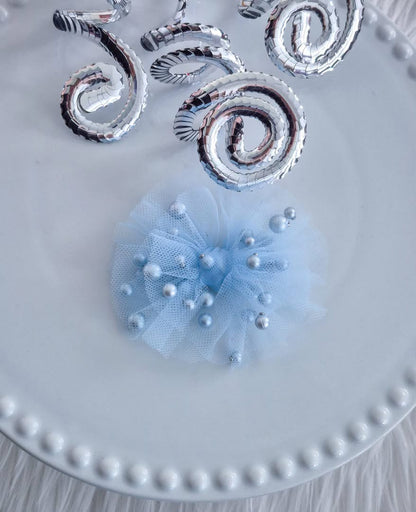 A light blue tulle hair bow embellished with scattered pearl beads, displayed on a white beaded-edge plate with metallic silver spiral decorations in the background.