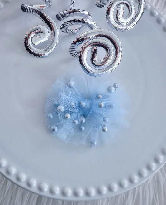 A light blue tulle hair bow embellished with scattered pearl beads, displayed on a white beaded-edge plate with metallic silver spiral decorations in the background.