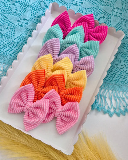 A set of six different small piggies set bows in assorted pastel and bright colors, arranged on a white scalloped tray over a turquoise lace cloth.